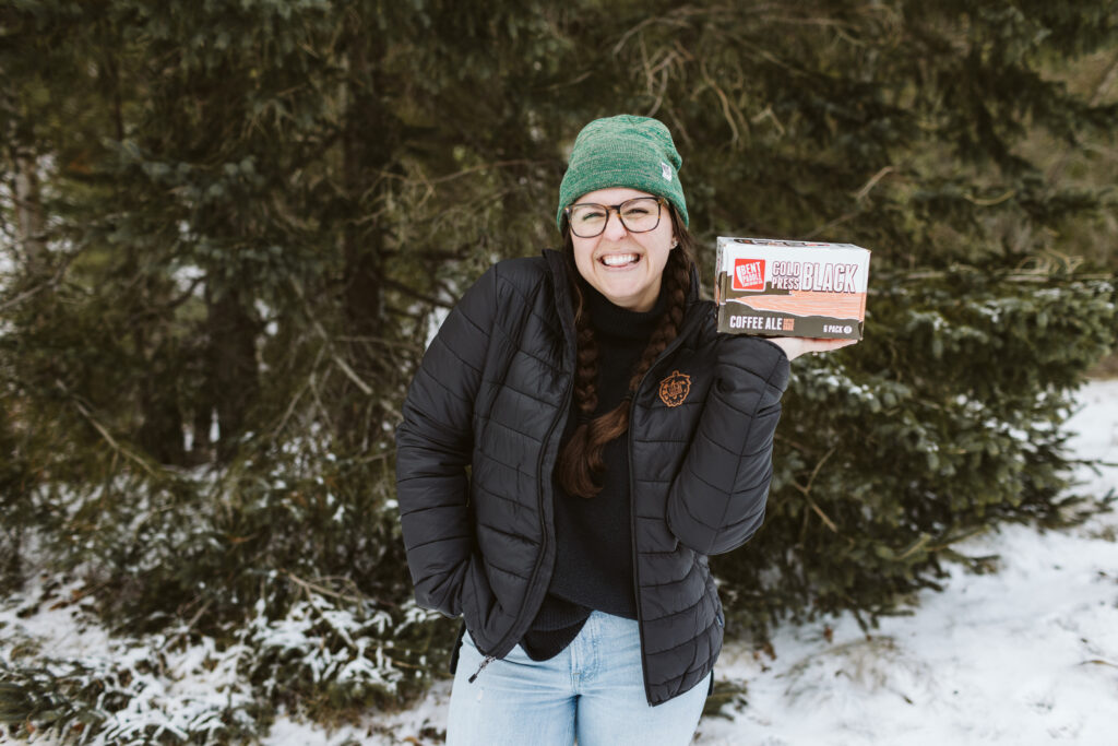 Women wearing bent paddle jacket and hat holding cold press black coffee ale 6-pack in snowy setting